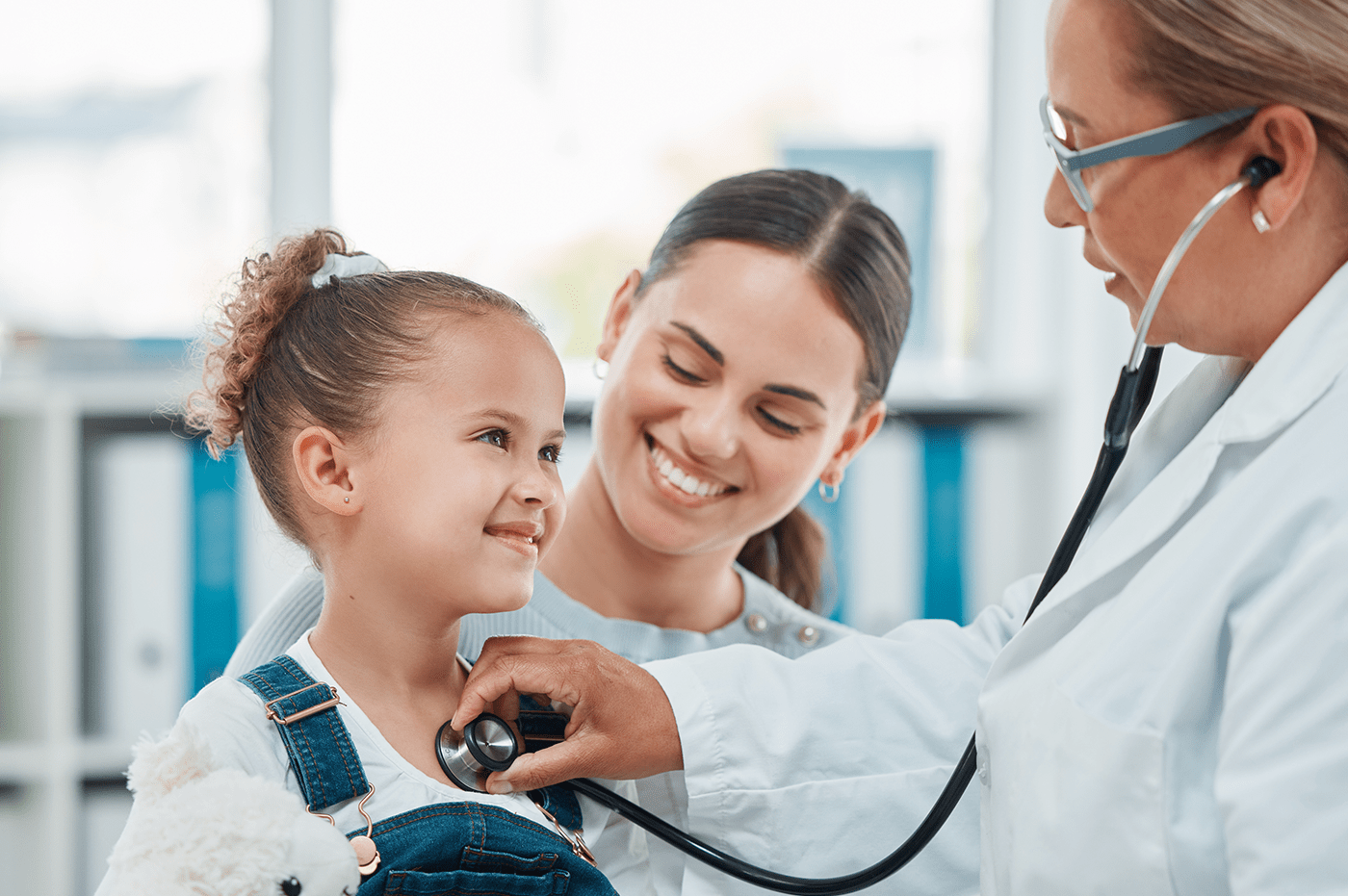 Doctor checking a little girl's heartbeat with her mom sitting with them.